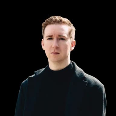 An image of Patrick McDonald, a white man in his 30s with brown hair wearing a black polo neck and grey wool coat.