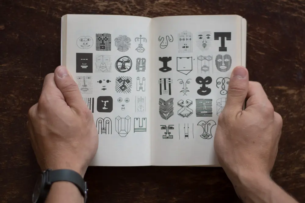 Two hands holding upon a double-page book spread. The page has many drawings of abstract faces of various styles.